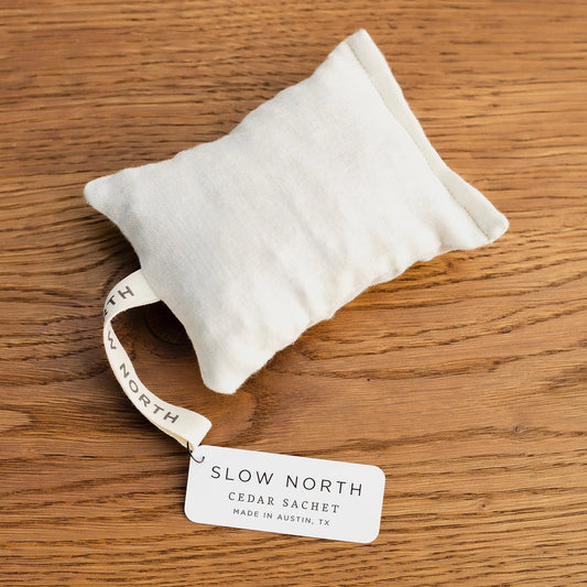 A Slow North Cedar Sachet resting on a wooden surface with a branded tag labeled "Best Seller," showcasing its natural cotton material and simple, rustic design.
