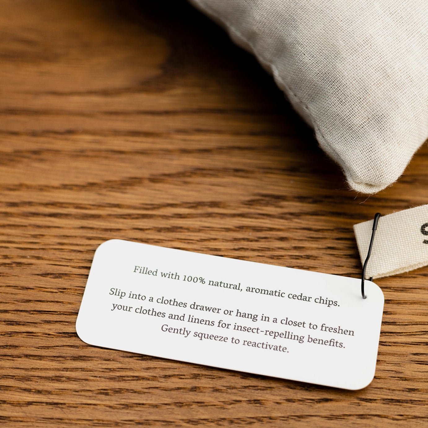 A close-up of the Slow North Cedar Sachet’s product tag, detailing care instructions and benefits, displayed on a wooden surface beside the natural cotton pouch.