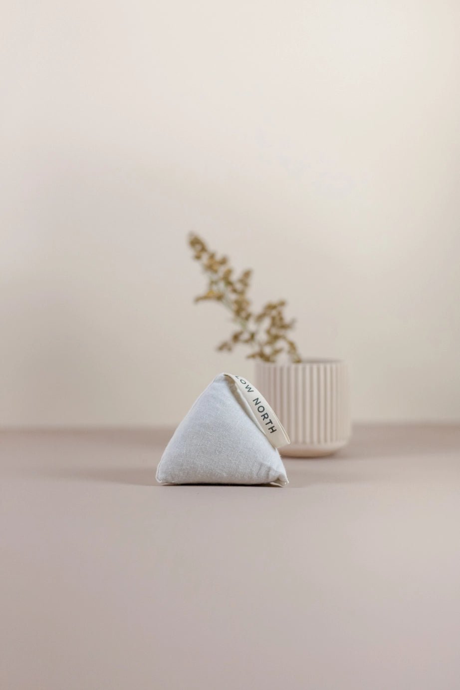 Single Slow North Cedar Sachet in a triangular form placed on a taupe surface with soft-focus dried florals in the background.
