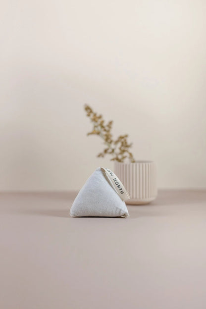 Single Slow North Cedar Sachet in a triangular form placed on a taupe surface with soft-focus dried florals in the background.