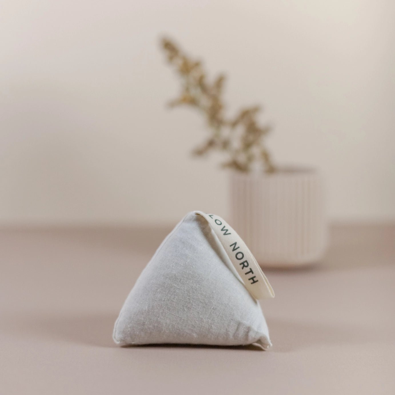 Slow North Cedar Sachet in a triangular shape standing upright on a neutral surface with a blurred vase and dried florals in the background.