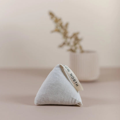 Slow North Cedar Sachet in a triangular shape standing upright on a neutral surface with a blurred vase and dried florals in the background.