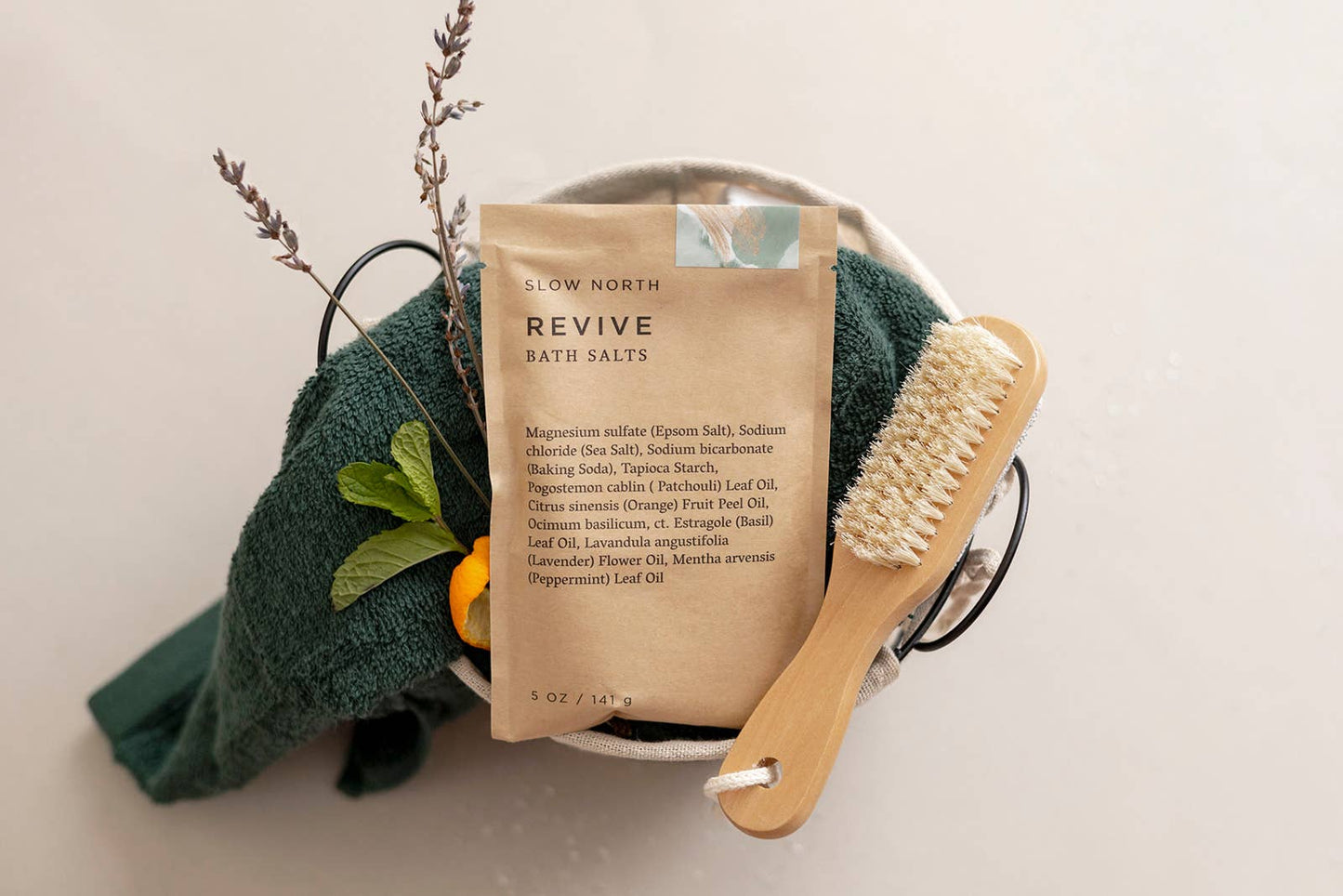 Slow North Revive Bath Salts displayed in a spa gift basket with a green towel, wooden dry brush, and lavender citrus accents.