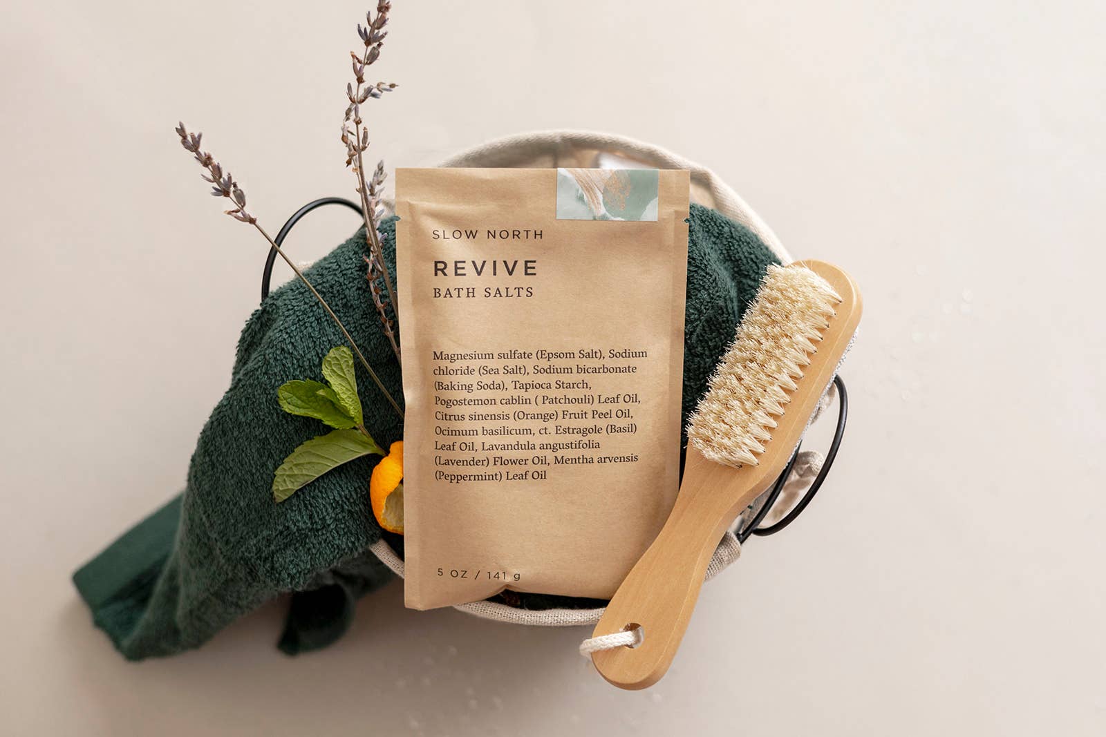Slow North Revive Bath Salts displayed in a spa gift basket with a green towel, wooden dry brush, and lavender citrus accents.