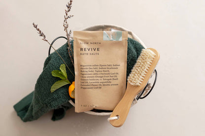Slow North Revive Bath Salts displayed in a spa gift basket with a green towel, wooden dry brush, and lavender citrus accents.