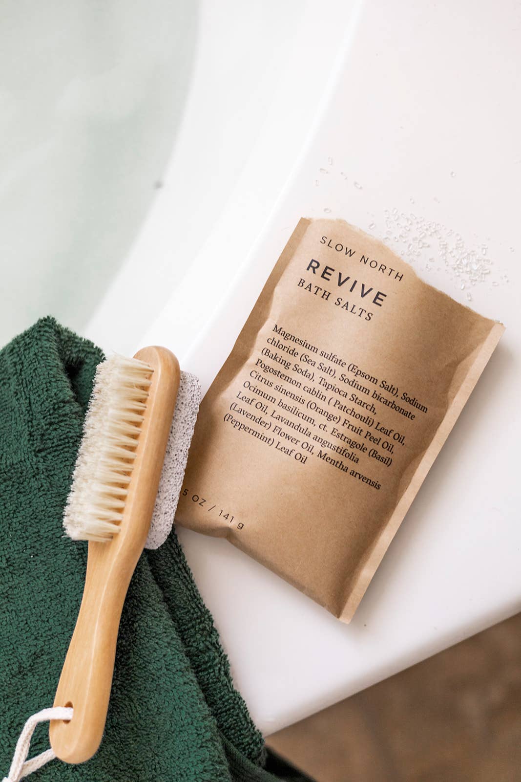Slow North Revive Bath Salts resting on the edge of a bathtub with a green towel and wooden bath brush nearby.