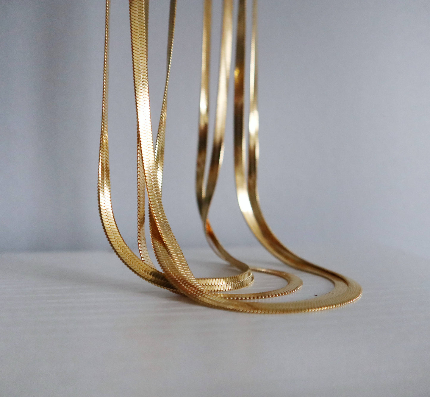 Close-up of multiple gold snake herringbone chain necklaces hanging and overlapping in warm light with an off white grey surface background.