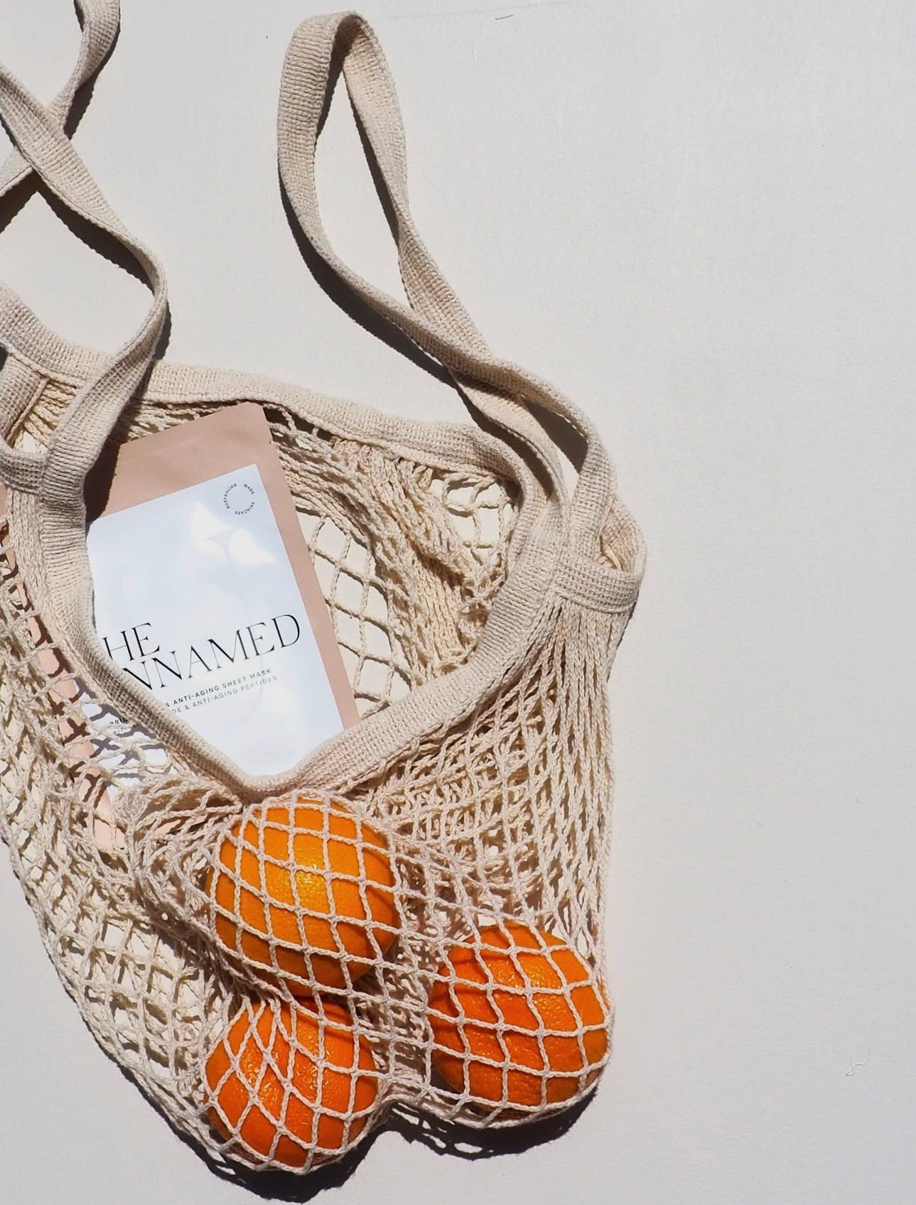 The Unnamed Brightening & Anti-Aging Sheet Mask tucked in a mesh market bag with oranges.